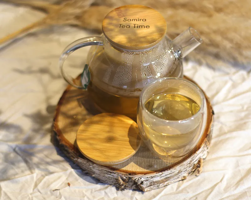 Théière en verre avec couvercle en bambou gravé, et une tasse de thé en verre transparent, posées sur un rondin de bois