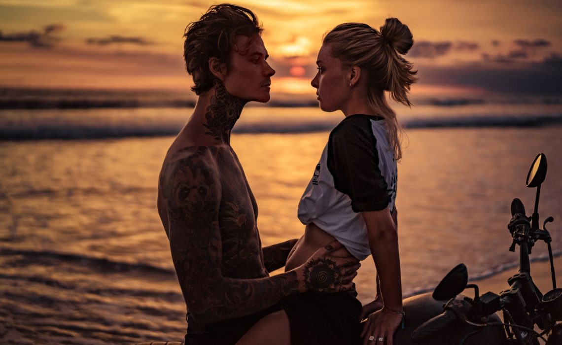 Un couple assis sur une moto se regardent avec désir devant le coucher de soleil