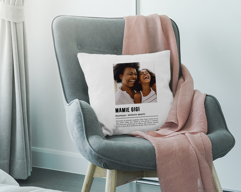 Cadeau personnalisé Fête des grands-mères 2025 : coussin 'Définition Mamie' sur fauteuil en osier avec plantes environnantes.