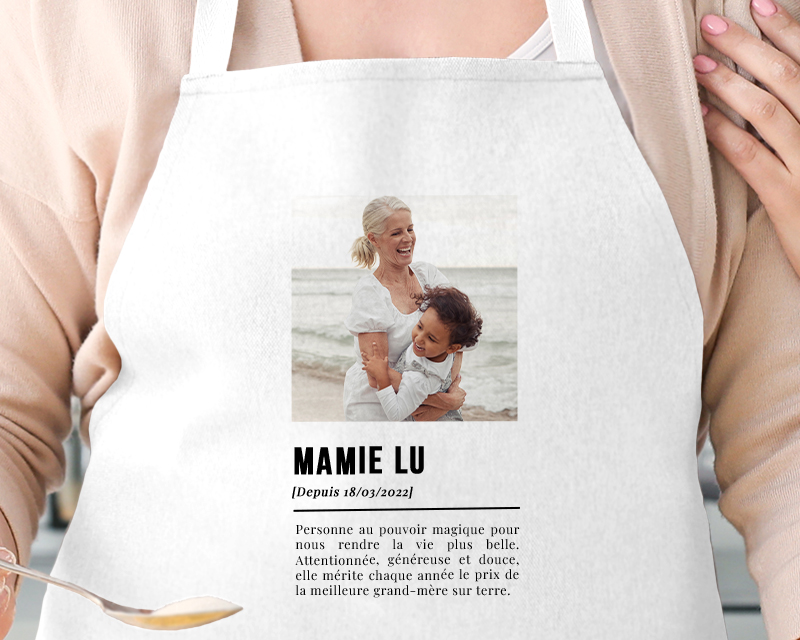 Cadeau Fête des mamies : tablier de cuisine blanc personnalisé avec photo et définition 'Mamie', porté par une grand-mère souriante en cuisine.